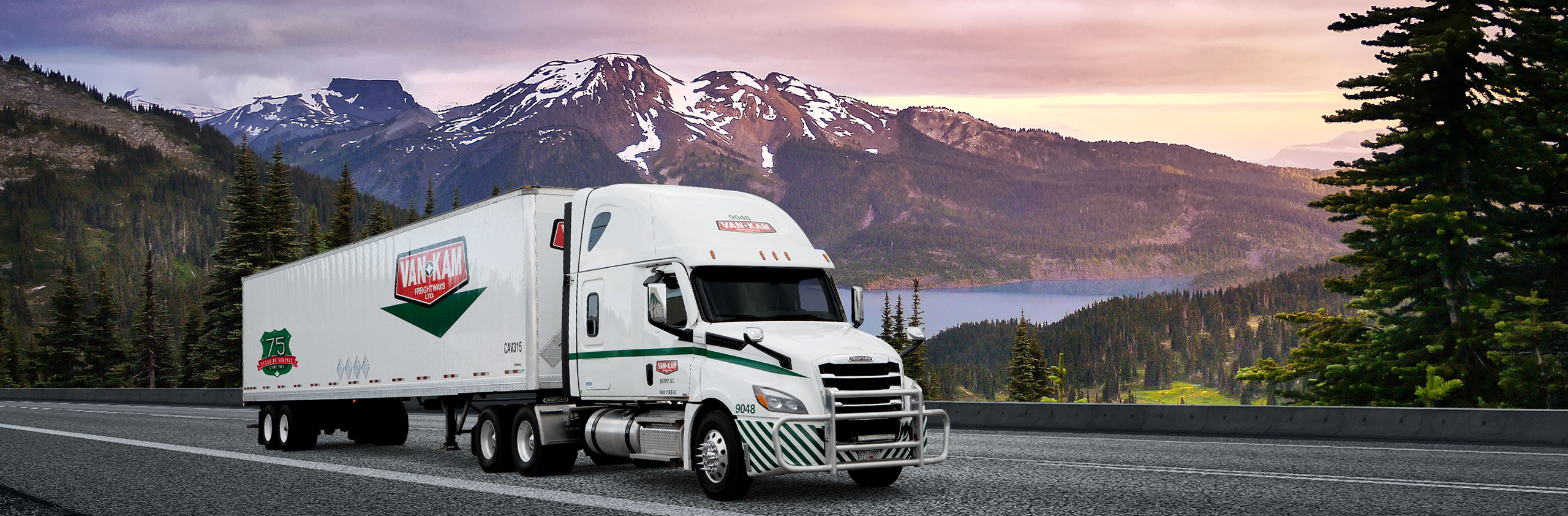Van Kam highway truck and 75th anniversary trailer driving in front of BC mountain landscape